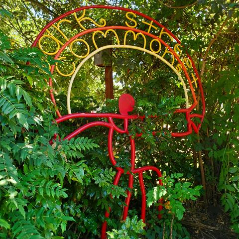 Red & yellow metal painted art, large scale, standing entry sign piece that says Wark Street Commons. It's surrounded by green plants foliage.