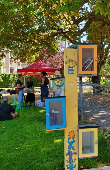 4 Little Free Library boxes, mounted on wooden post, painted in bright blue, green & yellow with some flowers & books painted on post & boxes. In background is event tent with creative instructors setting up for the workshop.