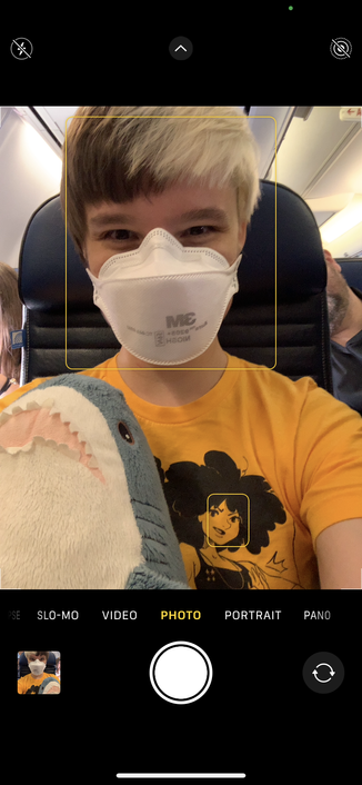 iOS camera interface shows a masked B holding a small IKEA Blahaj on an airplane. Their face is highlighted with a box showing the autofocus targets, and their shirt also has a face highlighted but the face is a cartoon drawing.