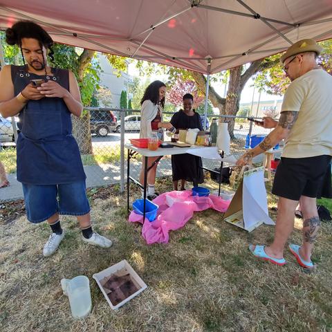 Kemi & an attendee working on mixing up coffee based photo developer at table. Another attendee is standing on the right. Joshua is standing on the left. A photo developing that with photo negatives in water is on the ground, with a pitcher of water, in front of Joshua's feet.