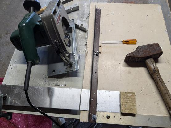Sur la table, une scie circulaire et un gabarit (une tôle plate à angle droit avec une cornière, les 2 vissés sur une planche en bois) permettant de couper des panneaux aluminium identiques