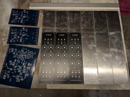 Sur la table, 12 panneaux en aluminium (71mm de large, hauteur 3U et 6U), à gauche 3 PCBs et 3 faces avants de VCOs