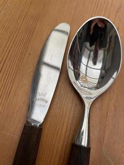 A butter knife and a spoon with wooden handles are placed on a wooden surface. The butter knife has the brand "SKAUGUM RUSTFRI" engraved on it.