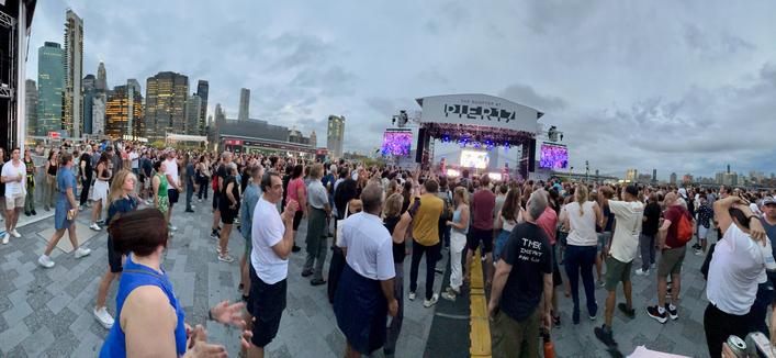 A panoramic view from the back of the floor at The Rooftop at Pier 17 in NYC at the Michael Franti & Spearhead show on Friday July 12, 2024. The Manhattan skyline is visible to the left, the stage is seen off center, the Brooklyn Bridge is to the right and a crowd of people are gathered on the floor.