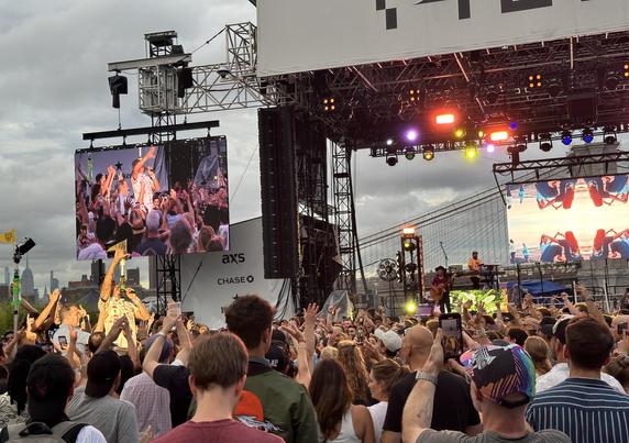 A view of Michael Franti at The Rooftop at Pier 17 in NYC at the Michael Franti & Spearhead show on Friday July 12, 2024. He is singing on a platform in the crowd on the floor, looking to the sky with his right hand in the air. A video screen & the stage is behind him.