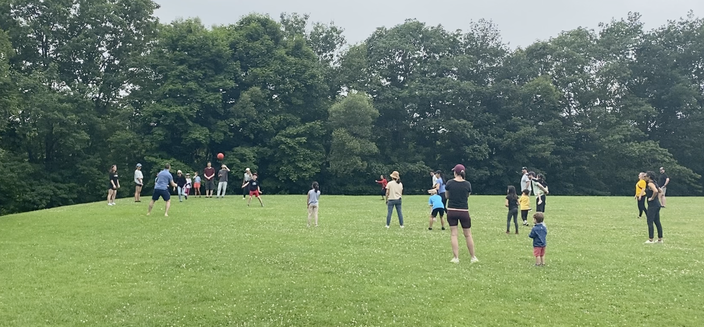 A group of adults and children on a field surrounded by trees playing kickball. The sky in the background is cloudy and there is a red ball being kicked into the air. Everyone is having fun!