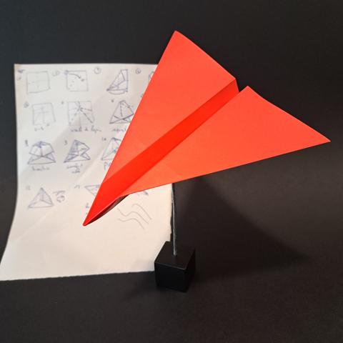 Un avion de papier rouge, en origami. Il est maintenu par une pince métallique. en arrière-plan, le diagramme du modèle, dessiné à la main sur un bout de feuille blanche.