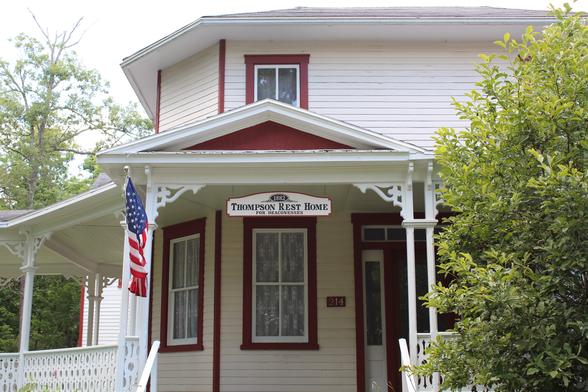 A white Victorian-era house with red trim. A sign hanging from the porch roof says "Thompson Rest Home for Deaconesses."