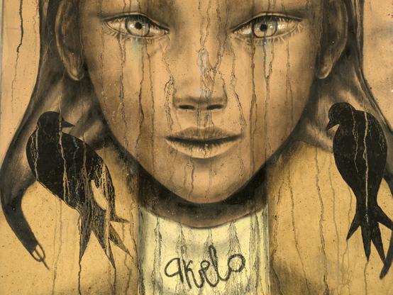 Fresque murale dans les tons sépia représentant une partie de visage d’une jeune fille avec des yeux larmoyants, comme si la peinture bleue et brune en coulait. Sur chaque épaule se trouve un oiseau noir. Sous le cou, le nom de l’artiste est inscrit.