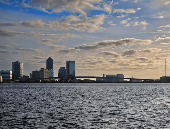 Beneath a sunny blue sky with large white and grey cloud formations a city skyline of colorful skyscrapers and bridges spanning a wide, choppy, blue river.
