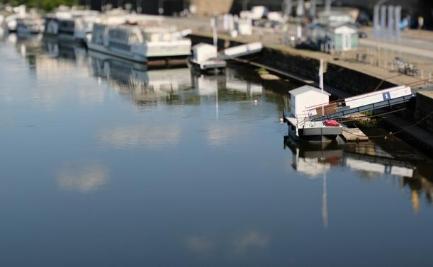 Fotografie von einem Bootsanleger an einem Fluss. Der Blick geht augenscheinlich von einer Brücke aus gegen die Flussrichtung des Gewässers. Am rechten Bildrand ist der Anleger, dessen Steg auf eine gepflasterte Straße führt. Im Hintergrund sind verschwommen Schiffe an ähnlichen Anlegern zu erkennen. Die ganze Szenerie hat aufgrund der sehr geringen Tiefenschärfe den Eindruck einer Miniatur.