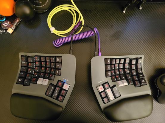 A Kinesis Advantage 360 keyboard sits on a desk with a carbon fiber pattern. The keyboard is a dark gray color, has black palm rests, and the keycaps have been replaced with a third-party set of keys that have Arasaka iconography and Japanese characters on them alongside the English letters. The keyboard halves are connected with a black cable, and the keyboard is connected to a docking station (out of frame) by a tightly-coiled cable that is a bright purple, which connects to a day-glo green extension cable.
