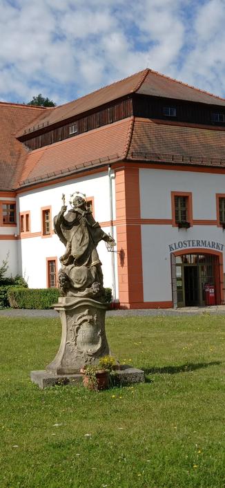 eine Heiligenfigur mit vergoldetem Heiligenschein steht auf einer Wiese vor dem Haus des Klostermarktes vom Kloster  Marienthal