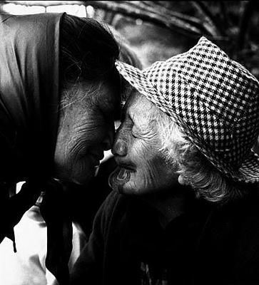The image depicts two older women sharing a warm and intimate moment. They are facing each other with their foreheads touching, suggesting a deep connection or affection between them. The woman on the left is wearing a dark-colored headscarf and has her hair pulled back, while the woman on the right is donning a light-colored hat that matches her attire. Their eyes are closed, emphasizing the tender nature of their interaction. The photograph is captured in black and white, which lends a timeless quality to the scene, focusing the viewer's attention on the emotional connection between the two subjects.