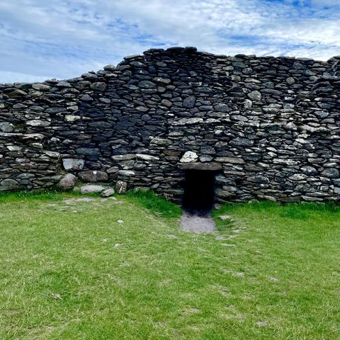 Ancient stone wall with a small, dark entrance at the base and green grass in the foreground. Blue sky with clouds in the background.