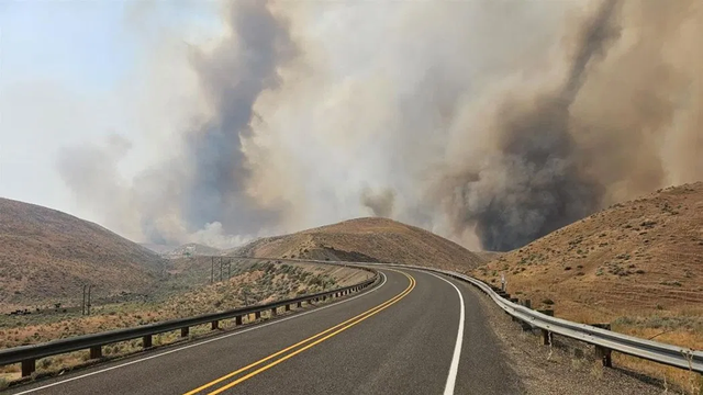3 huge grey and black plumes of smoke  rise above 2 lane  highway between two valleys