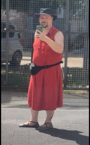 Ein Mann mittleren Alters (ich) und kräftiger Statur steht in einer Fußgängerzone. Ich trage ein wadenlanges, ärmelloses Leinenkleid mit Hemdkragen und ausgestelltem Rockteil. Die Farbe ist Terracotta-Rot. Dazu einen schwarzen Hut, eine schwarze Bauchtasche und graue Ballerinas mit Kreuzgummis.