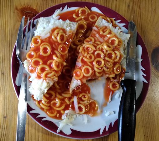 Close up if a white plate with burgundy pattern around the edge with a fork on the left and a black handled knife on the right. The meal is two pieces of bread covered in pale yellow grated cheese and covered in spaghetti hoops in tomato sauce.