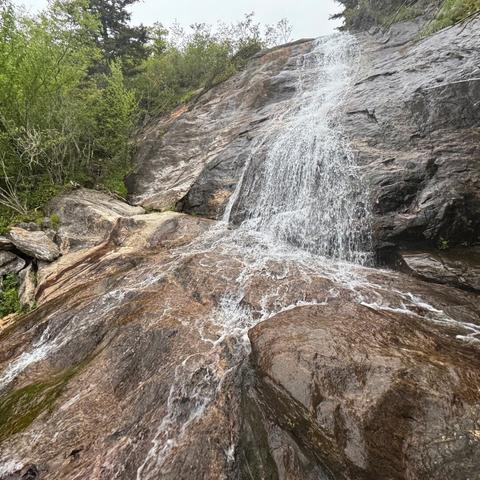 Graveyard Fields Upper Falls in North Carolina. A thin stream of water cascades down a tall rough surface of rock, some of which is dark but lower bits are lighter with an orange tint.