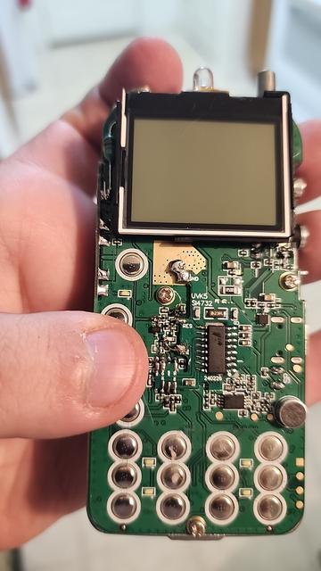 A Quansheng UV-K5 handheld radio with the cover removed, showing the circuit board. A green resistor has been added just beside the display, an additional daugher board has been soldered on with a haphazard looking blob at the top (ground connection), and a series of solder jumpers from the existing board to the daughter board on top of it. There's even one solder connection that required a miniscule piece of copper wire to make the connection. The 16-pin DIP IC in the center of the daughter board isn't even as long as my thumb is wide.