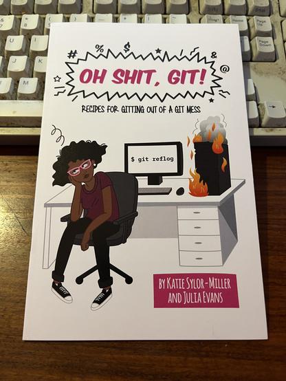 A copy of the dead-tree version of "Oh shit, Git!" by Katie Sylor-Miller and Julia Evans. The cover shows a typical day in the office with a developer whose head is about to explode because git has set her computer on fire.
