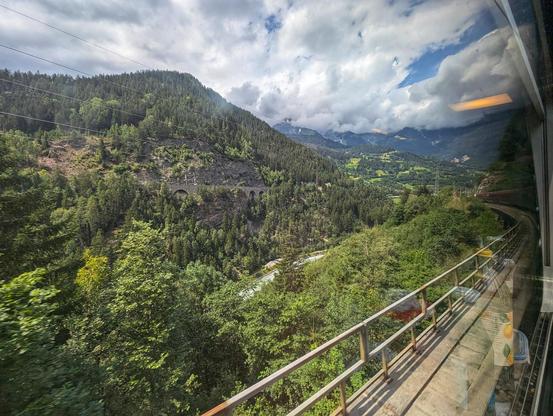 Blick auf den Inn und zurück auf den Zug am Arlberg mit den Hügeln und Gewölk. Am Gegenhang zeigt sich ein Lehnenviadukt als gemauerte Naturstein-Bogenbrücke.
