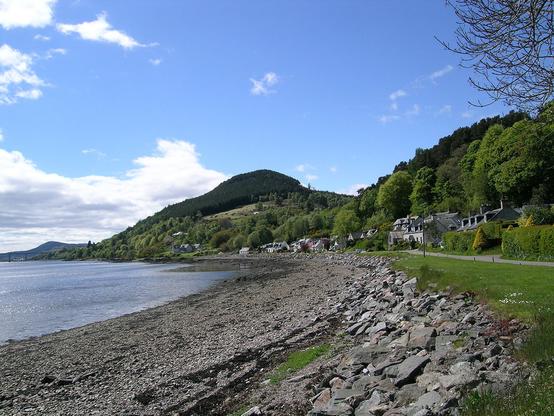 Kilmuir. The image shows a view along a rocky beach that curves to the left and has sea to its left. There is grass above the beach and a road, and to the right and above the road is a line of houses and cottages that blends back into a hillside covered in trees. The sky is largely blue.