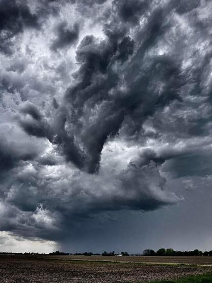 Photo of some storm clouds over Oswego, Illinois. While the image is in color, it seems almost black and white. Among the thick gray clouds is a very dark black cloud, which is beginning to take the shape of a funnel cloud. It is twisted into multiple columns.

Below is a large swath of open farmland and prairie.

[Photo credit: Paul Rubicki ]