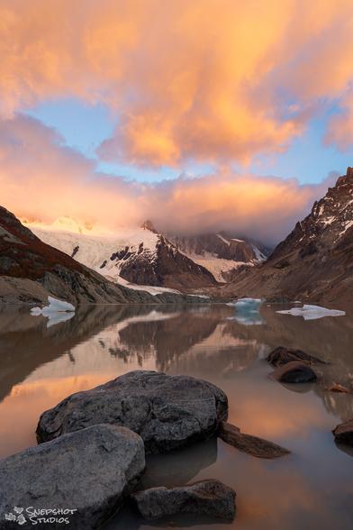 A lake in-between mountains. Some rocks in the water in the foreground. Snowfields and glaciers behind the lake. Brightly lit orange clouds in the sky above.