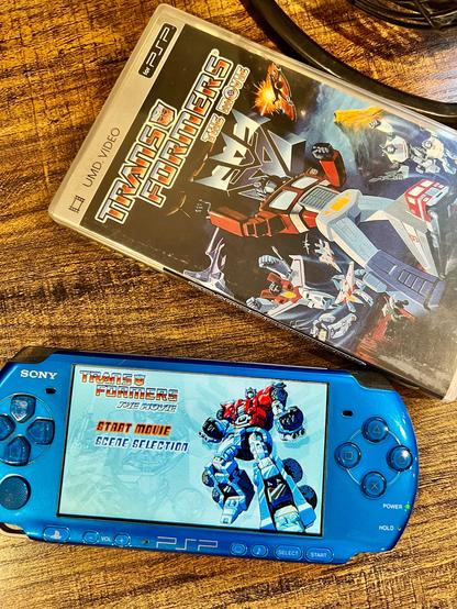 A photo of a blue PSP showing the menu screen for the UMD Video version of Transformers The Movie. Above the PSP is the case for the movie.