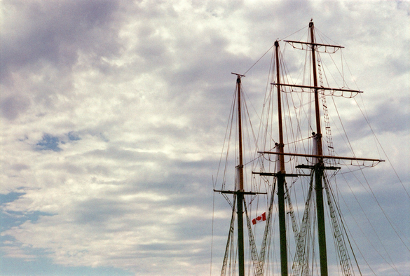 The upper two thirds of the masts of a tall ship against a mostly cloudy sky.