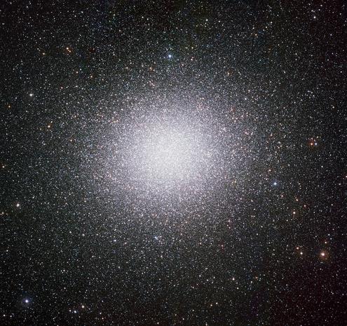a bright, ball of stars, collected together centrally. The cluster of stars features mostly white stars but there are numerous blue and red stars also. The density of stars is highest at the centre, and drops radially, giving the perspective of a hive-like structure swarming with points of light.