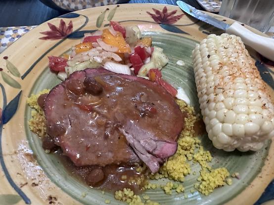 A plate of food featuring a slice of roast beef with gravy, a serving of couscous, a mixed vegetable salad, and a seasoned ear of corn.