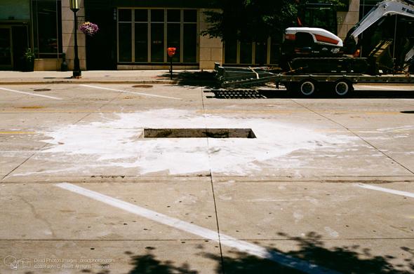 A photograph of a rectangular hole in the center of a concrete street. The hole has smooth edges, and the pavement around is in all directions is covered in a bright white mud. There are painted white diagonal parking lines on both sides of the street, which is empty except for a white and red Bobcat backhoe on a flatbed trailer. It is sunny, and the trailer and street-side trees cast dark shadows.