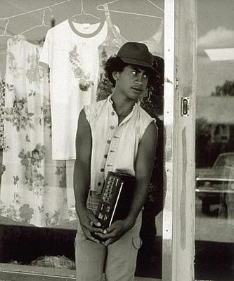 In the image, a young man is standing in front of a shop window display. He appears to be looking into the window with interest. The man has curly hair and is wearing a hat, which adds to his casual appearance. His expression seems contemplative or curious about what he's observing.

The setting suggests it might be a sunny day outside the shop. There are other items in the display behind him that could potentially interest a shopper, but they are not clearly visible due to the focus being on the man in front of the window. The style of the photograph and its composition give the image an artistic quality, suggesting it might be a piece of photographic art or used for commercial purposes related to fashion or retail.