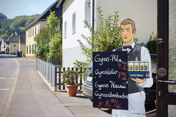 deutsch: das querformat zeigt eine impression aus dem eifeldorf urft zwischen kall und nettersheim.
links zu sehen ist ein gastronomieaufsteller in form eines kellners, der einen kranz kölsch in der linken hand hält, davor eine schwarze fläche, auf der vier verschiedene gyros-variationen mit den entsprechenden preisen angegeben sind.
von der unteren bildmitte nach links oben ist die hauptstraße des ortes, die urfttalstraße und der gehweg zu sehen. ganz im hintergrund bewaldete hügel.

english: the landscape format shows an impression of the eifel village of urft between kall and nettersheim.
on the left is a catering stand in the form of a waiter holding a wreath of kölsch in his left hand, in front of it a black surface on which four different variations of gyros are indicated with the corresponding prices.
from the bottom centre of the picture to the top left you can see the main street of the village, urfttalstraße and the footpath. in the far background are wooded hills.