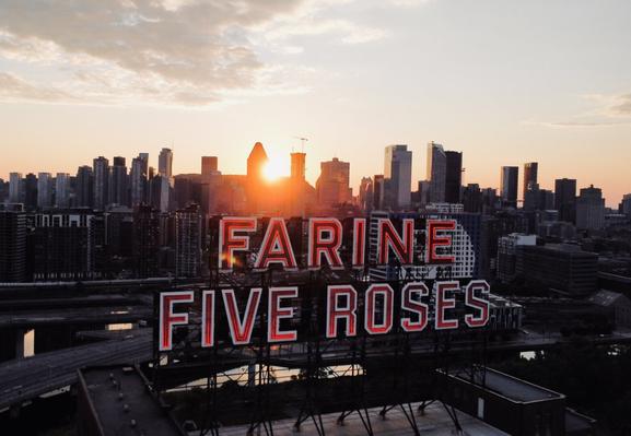 A photo taken in Montreal at sunset, showing the iconic Farine Five Roses sign with the city's skyline in the background. The sun is setting behind the buildings, casting a warm glow over the scene.