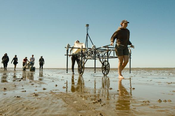 Mehrere Personen gehen durchs Wattenmeer und ziehen bzw. schieben dabei Wagen. Auf dem vorderen Wagen sind deutlich Antennen und Messgeräte zu erkennen.