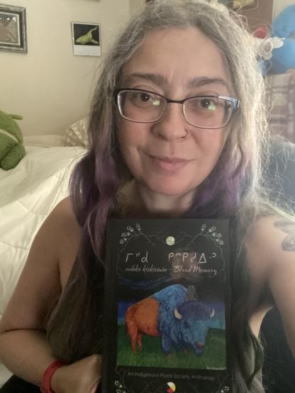 A person with long, graying hair and glasses holds a book titled "mihko kîsikâwin - Blood Memory: An Indigenous Poets Society Anthology" in front of them. The cover of the book features an illustration of a bison.