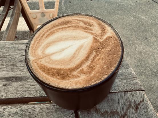 A small black coffee mug sits on the edge of a patio table with a frothy cappuccino with some simple latte art.