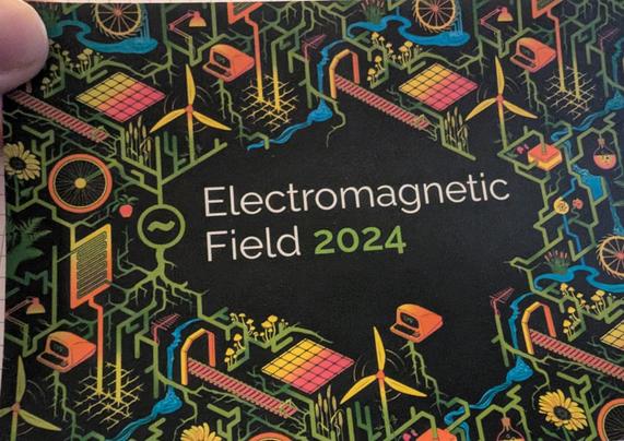 Postkarte des EMF24 mit Beschriftung "Electromagnetic Field 2024" auf schwarzem Grund umgeben von den Graphics der Veranstaltung: ein Wimmelbild "Solarpunk Network" aus stilisierte Pflanzen und Wurzelranken, Schienengleisen, Pilzen, Windrädern, PCs der jüngeren Generation, Rädern etc. und dem Logo des EMF in Farbtönen von grün über gelb-orange zu blau. Die bunten Farben leuchten stark LED-artig aus der Kartenebene. Räumliche Strukturen werden durch gerasterte Linien angedeutet, sodass einzelne Objekte aus der Ebene zu treten scheinen.