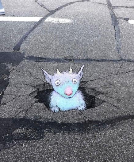 Streetart. On an asphalt floor with tar patches and cracks, a little blue furry monster called Kevin and a hole was drawn in chalk. It has pointy fluffy ears, two small horns, big eyes, a few nose wrinkles and a big pink nose. It looks out of its hole, a little confused. Title: "Kevin dug a hole all the way through the crust of the earth and then completely forgot what he came up here for..."