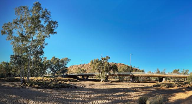 A bridge crosses a dry, orange-sanded riverbed, surrounded by gumtrees under a clear blue sky