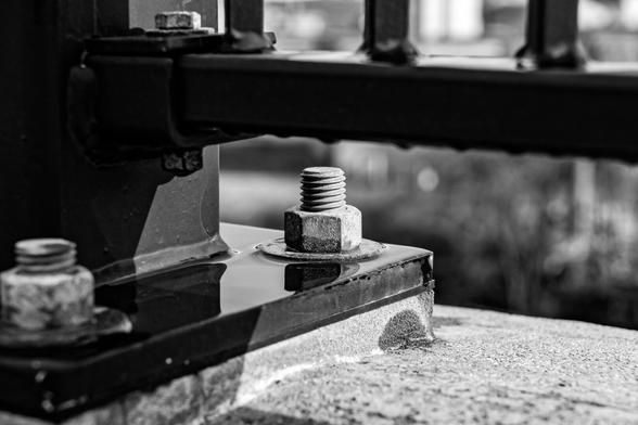 A black and white photograph showing the detail on a steel bolt holding a black metal safety barrier in place on a bike and pedestrian bridge 40+ feet over a river. The image has very shallow depth of field.