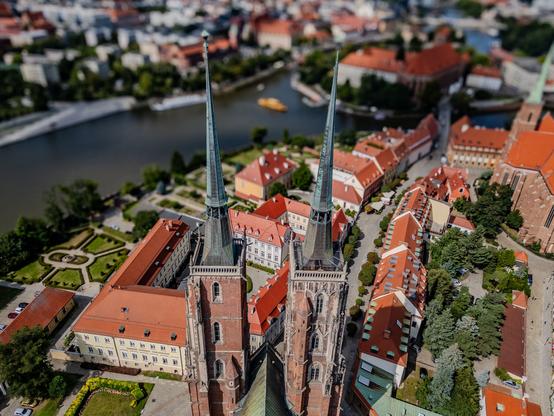 Aerial view of a European cityscape featuring two tall church spires, red-roofed buildings, green spaces, and a river running through the background.

Widok z lotu ptaka na europejski pejzaż miasta z dwiema wysokimi wieżami kościelnymi, budynkami z czerwonymi dachami, terenami zielonymi i rzeką przepływającą w tle.