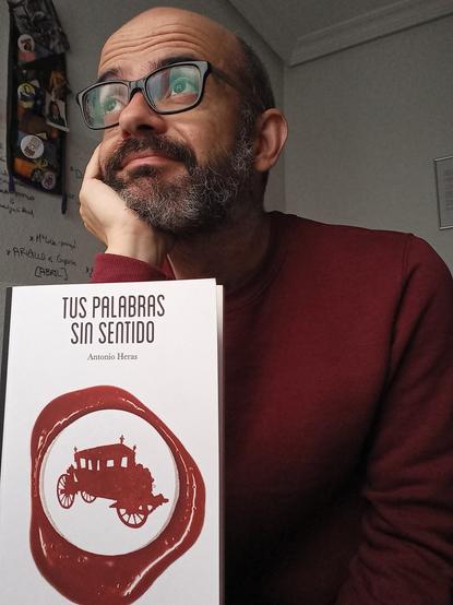 barbudo calvo con gafas mira al infinito con jersey rojo, delante libro puesto en pie, de fondo pared, fotos, chapas