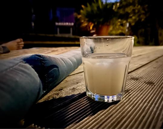 Glas Pastis neben einer Kapok Matte auf einer Holz Terrasse. Abends. Dunkel, aber ein beleuchteter Fokus im hinteren Teil.