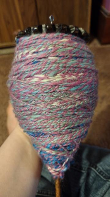 Drop spindle covered in lots of 2 ply multicolor yarn, completely dwarfing my adult female hand