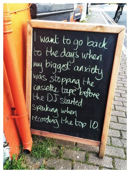A chalk written blackboard sign on the street. The handwritten text reads “I want to go back to the day's when my biggest anxiety was stopping the cassette tape before the DJ started Speaking when recording the top 10.”
