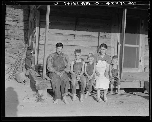 In this vintage photograph, a family of six is captured in their day-to-day life on a farm. The parents are seated in the middle, flanked by their four children. They appear to be dressed in work clothing, indicative of their role as sharecroppers or former mill workers from Gastonia textile mills who transitioned to farming after mill closures seven years ago. The family is situated on a dirt road next to a rustic wooden house, which serves as their dwelling. The overall scene depicts rural life with a touch of historical context.